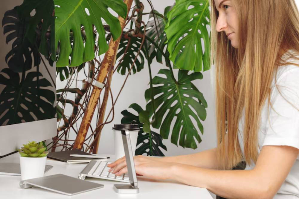 A woman types on a keyboard at a desk surrounded by large green plants, with a computer monitor and coffee cup nearby.