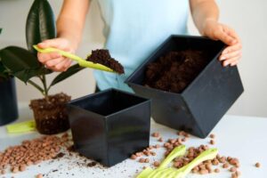 A person fills a black planter with soil using a green scoop, surrounded by scattered pebbles and a potted plant in the background.