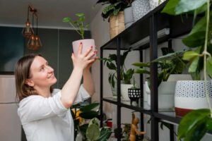 A woman in a white shirt holds a potted plant, surrounded by various green plants on shelves in a modern apartment.