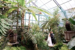 A couple in formal wedding attire stands amidst lush tropical plants in a greenhouse, surrounded by vibrant greenery and natural light.