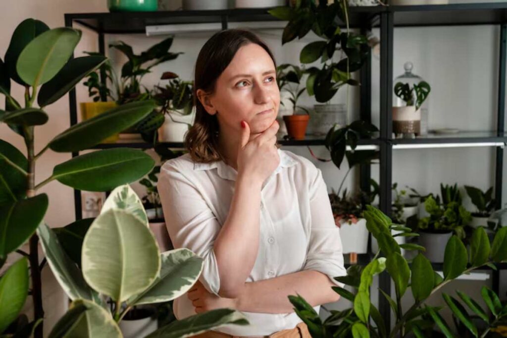 A person in a white shirt stands thoughtfully among vibrant indoor plants, with shelves filled with various greenery in the background.
