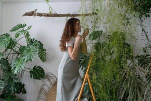 A woman in a light dress stands on a wooden ladder, surrounded by lush green plants, interacting with a hanging vine in a bright interior.