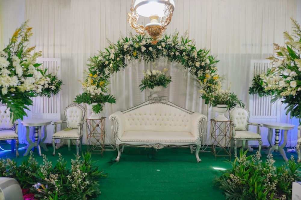 Elegant event setup featuring a white tufted sofa under a floral arch, surrounded by lush greenery and decorative tables.