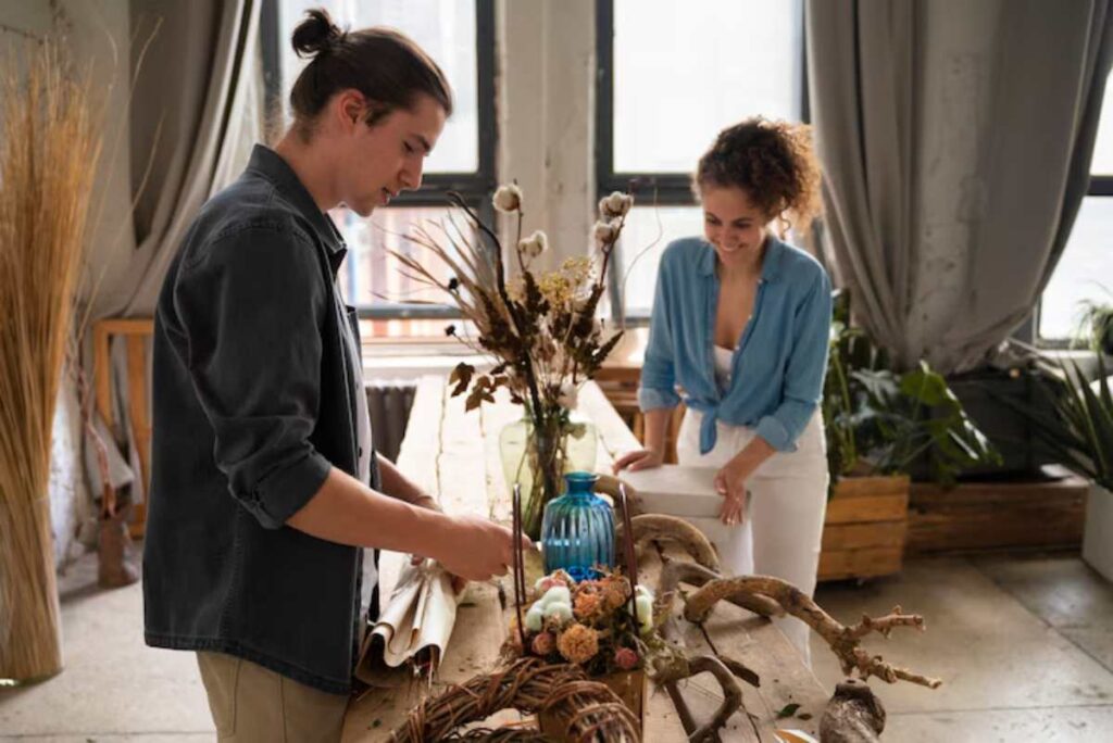 Two people are arranging decorative materials on a wooden table, surrounded by plants in a bright, stylish interior setting.