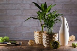 Two potted plants in woven baskets, a white jug nearby, and decorative spheres on a rustic table against a brick backdrop.