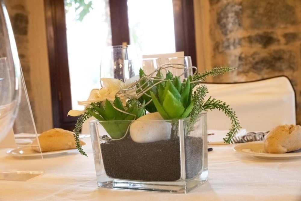 A glass vase with a succulent centerpiece filled with dark sand, white stones, and decorative greenery on an elegantly set dining table.