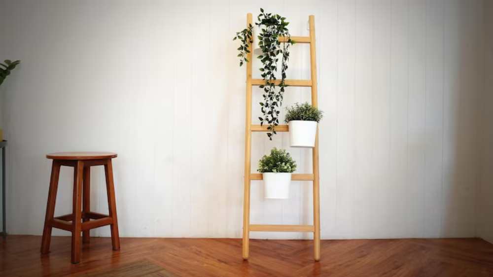 A wooden ladder shelf adorned with hanging plants in white pots, set against a plain white wall and beside a small wooden stool.