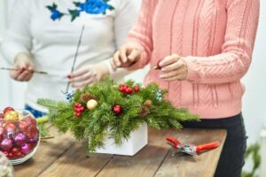Two women create a festive floral arrangement with pine, berries, and ornaments, working together at a wooden table.