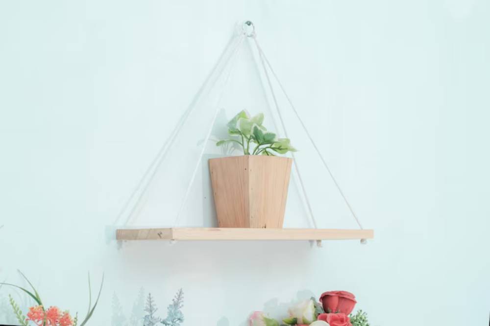 A minimalist wooden shelf suspended by ropes, featuring a potted green plant and colorful artificial flowers underneath.