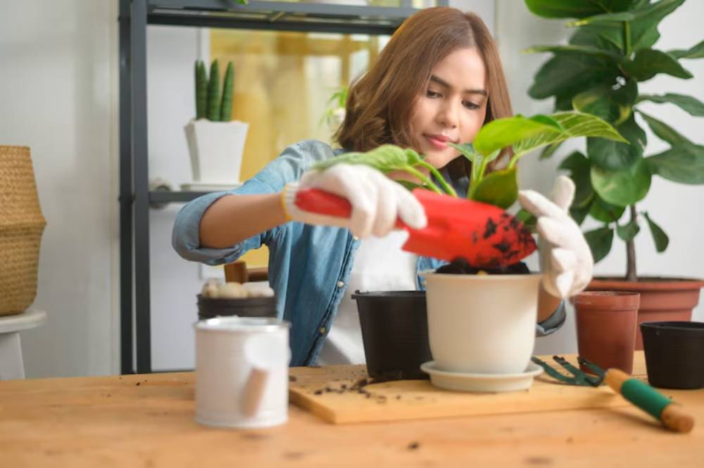 A person in gloves repotting a plant using a red trowel, surrounded by various pots and gardening tools on a wooden table.