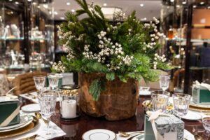 A festive table setting featuring a rustic wooden planter filled with greenery and white berries, surrounded by elegant glassware and gifts.