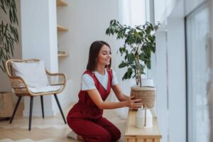 A person in a red jumpsuit kneels beside a wooden table, arranging a potted plant by a large window with natural light streaming in.